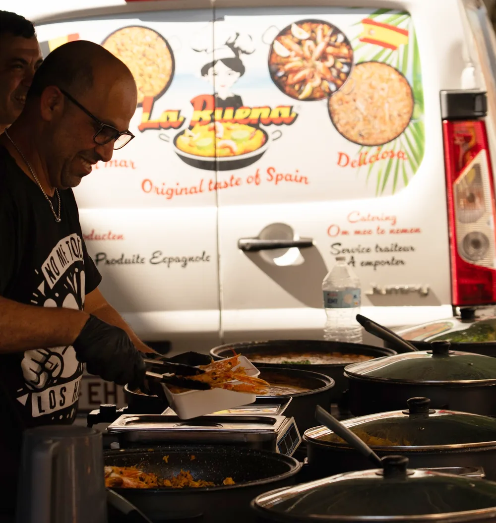 Een portie paella wordt opgeschept aan onze marktkraam in Leuven, met de bestelwagen van La Buena op de achtergrond